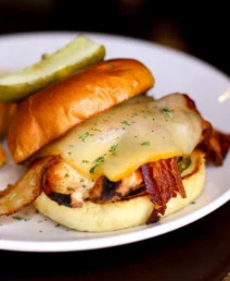 Gourmet chicken burger with bacon, cheese, and pickle on a white plate.